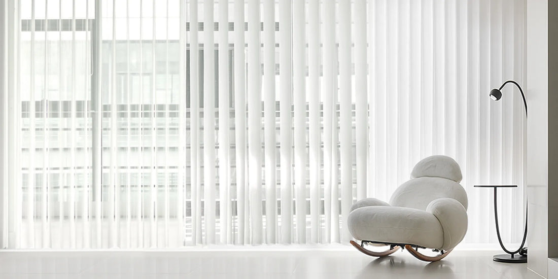 A modern white rocking chair in a minimalist room with sheer white vertical blinds covering a large window, alongside a black floor lamp and a small side table.