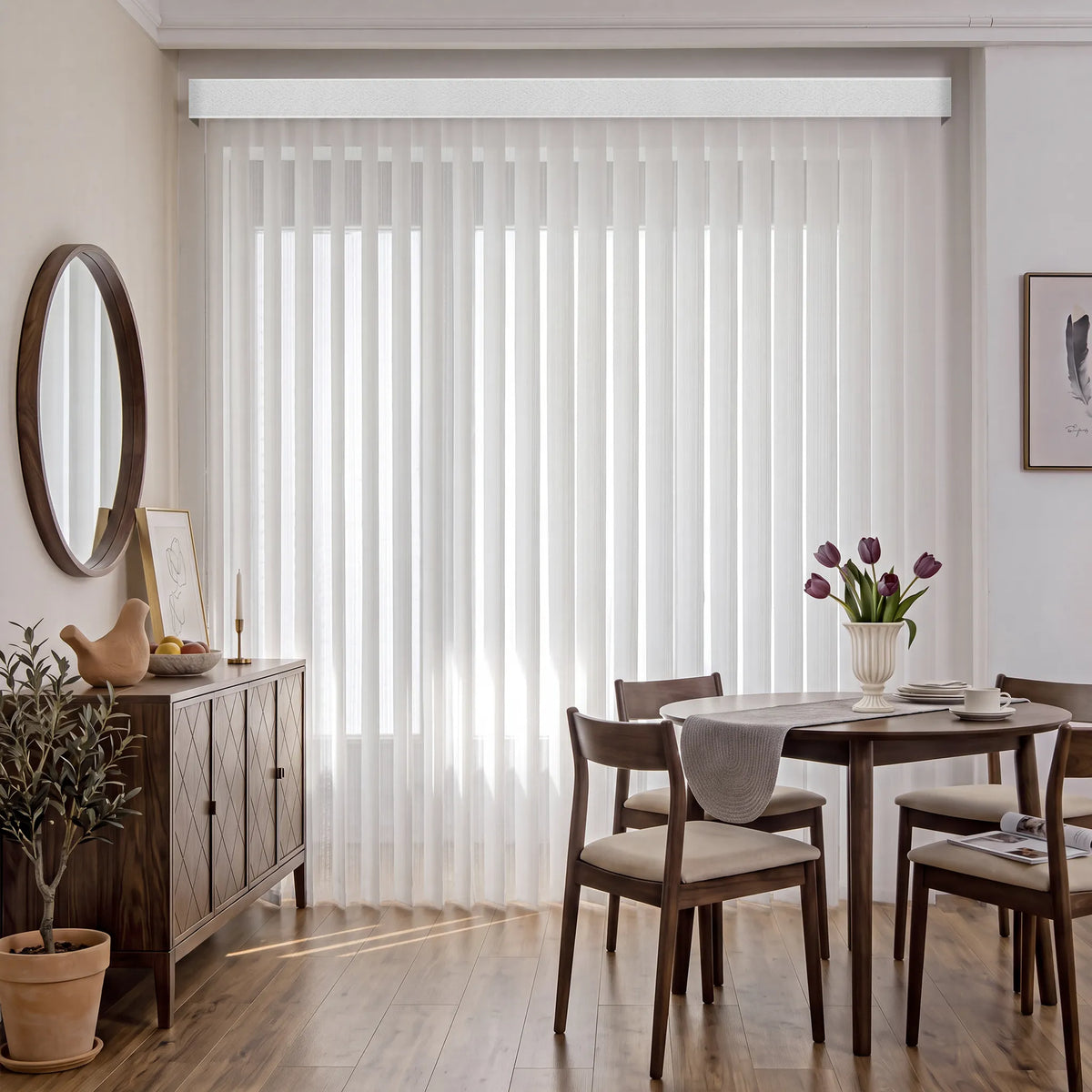 A dining room scene with a large window covered by white vertical blinds and a matching white Smart Sheer Vertical Blinds. The room includes a wooden sideboard, round wall mirror, dining table, chairs, vase of tulips, and light wood flooring.