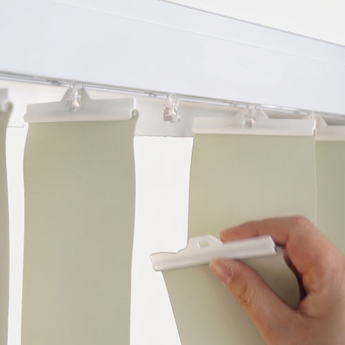 A close-up of a hand holding a white plastic clip to adjust or remove a light beige curtain panel hanging from a white rod with clear clips.