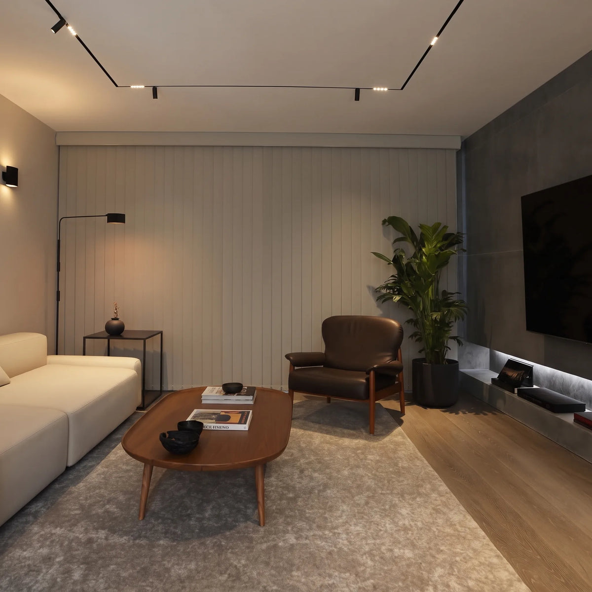 A modern living room interior featuring a white sofa, a brown leather armchair, a curved wooden coffee table, a large gray area rug, a black floor lamp, and a potted green plant against a light wood-paneled wall.
