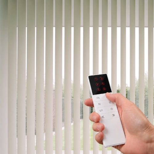 A hand holding a white remote control with a black touch panel, positioned in front of Smart Vertical Blinds covering a window.