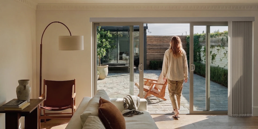 A woman stands with her back to the camera, walking through large sliding glass doors into a bright patio area. The scene is set in a modern, sunlit living room with a white sofa, a brown leather chair, a floor lamp, and a tiled outdoor patio.