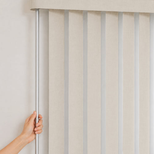 A hand holding a silver metal rod next to a beige vertical blind with light gray slats, set against a plain off-white wall.