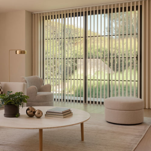 A modern living room with beige vertical blinds covering large floor-to-ceiling windows, with a light wood coffee table, upholstered armchair, round ottoman, and a view of a green outdoor garden.