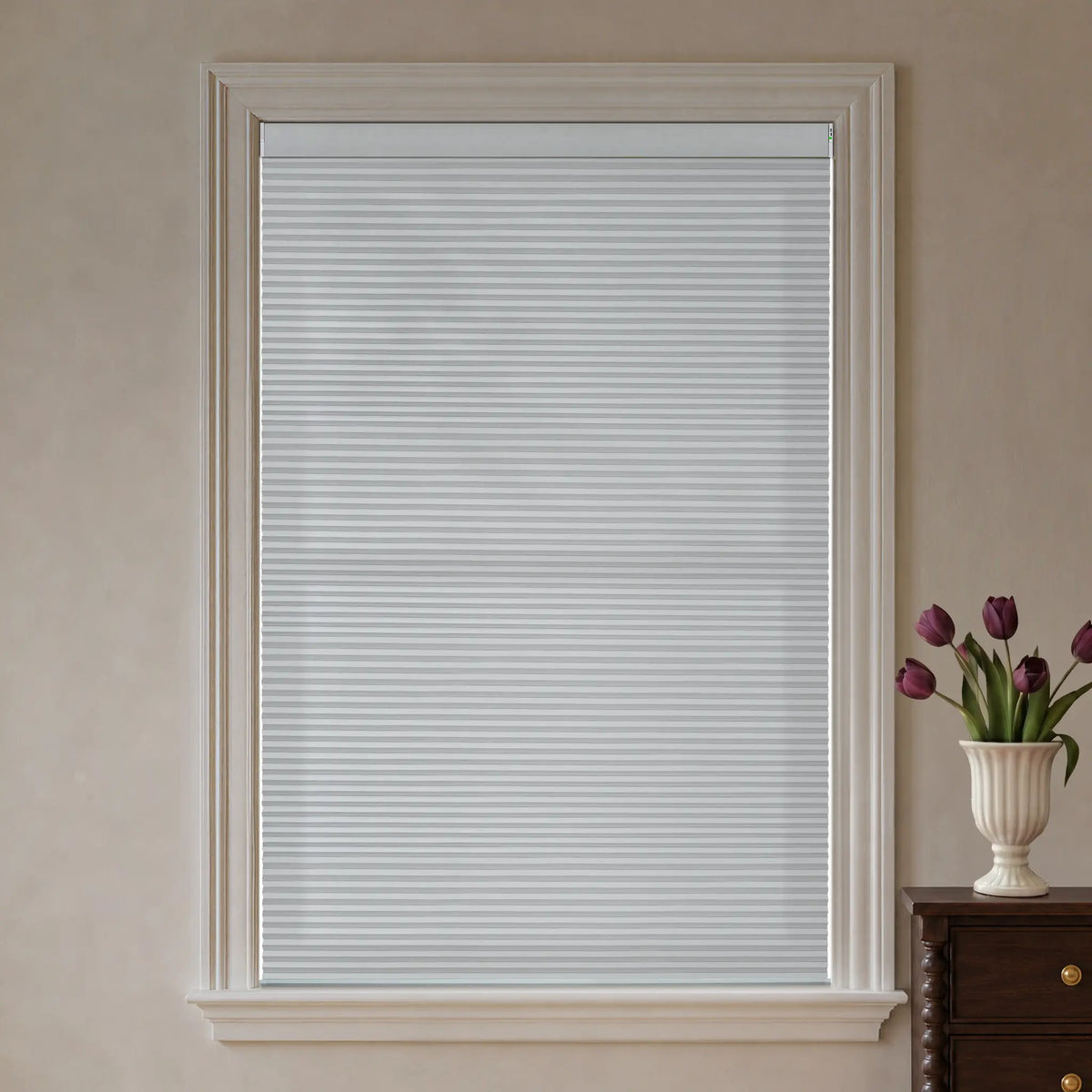 White motorized  cellular window blinds installed in a tall window with a white frame, set against a beige wall with a small wooden cabinet and a vase of purple tulips visible on the right.