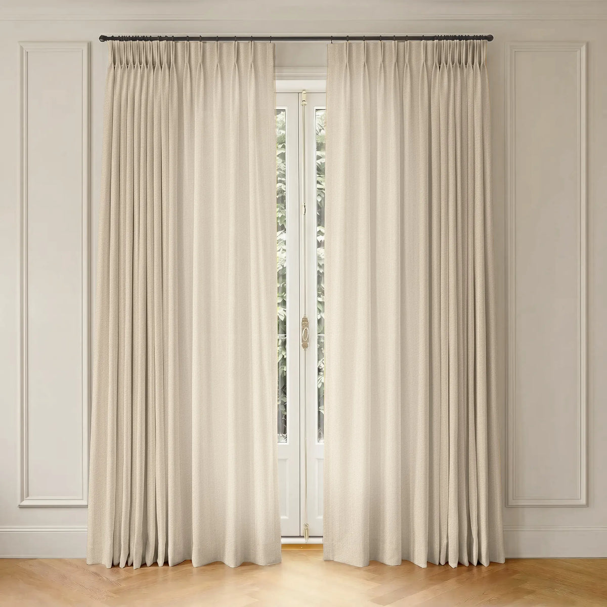 A pair of long cream-colored pleated window curtains hanging from a dark metal rod in a bright room with white paneled walls and a light wood floor.