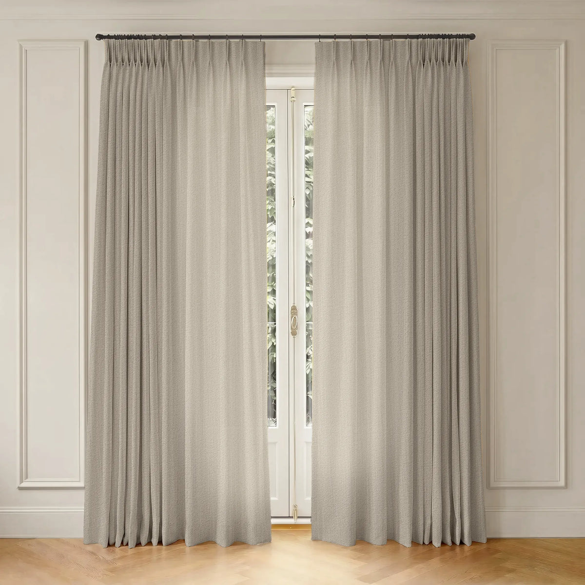 A pair of light beige textured window curtains hanging from a dark metal rod in a classic room setting with white paneled walls and a light wood floor.