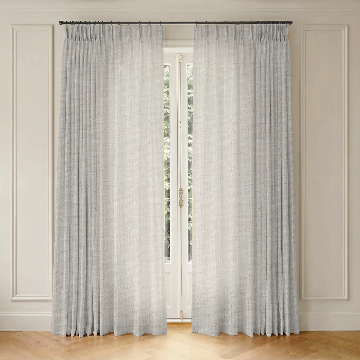 A pair of floor-length pleated curtains in a light gray textured fabric hanging from a dark metal rod over a tall window in a cream-colored room with wood floors.