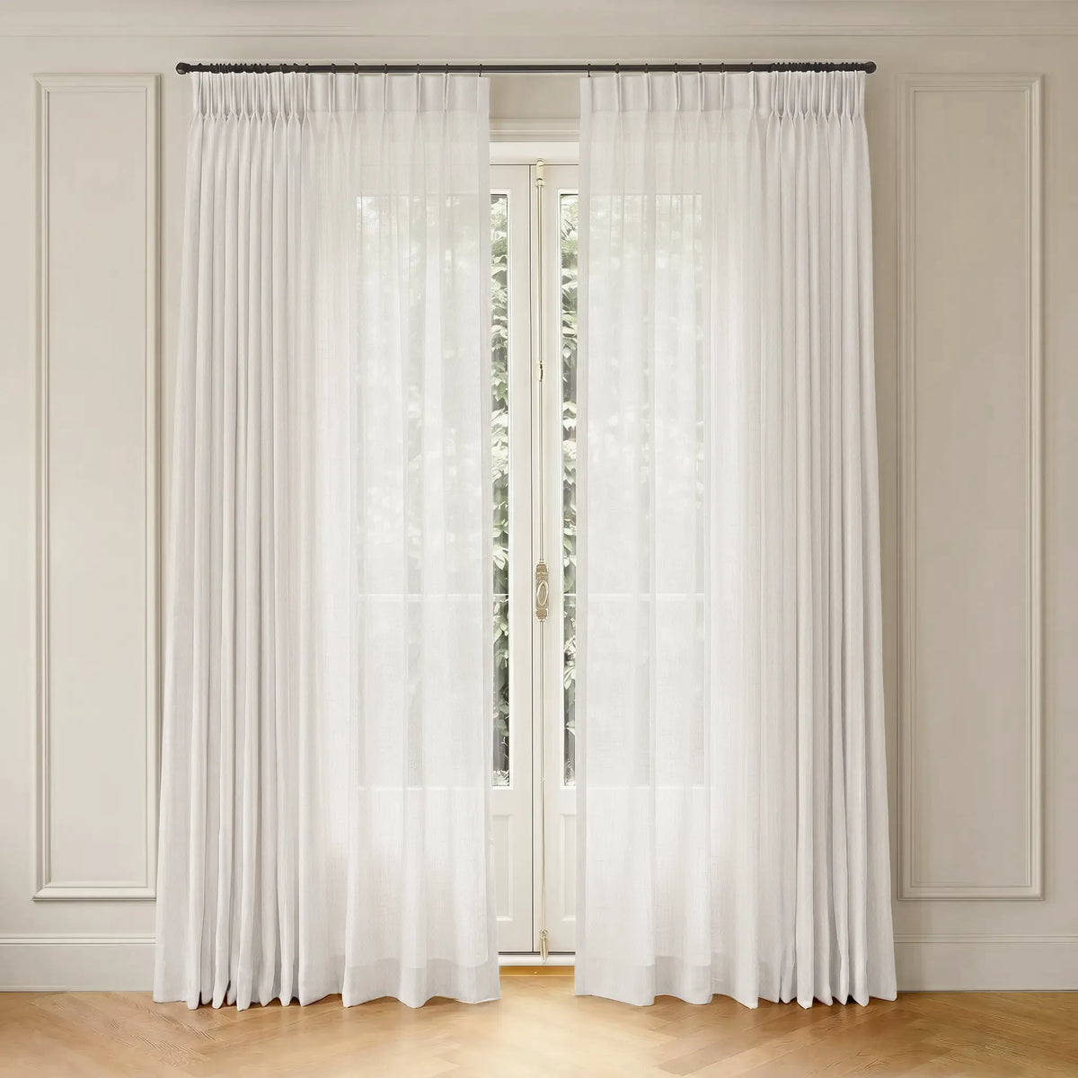 A pair of floor-length white sheer curtains hanging from a dark metal rod in a room with cream paneled walls and light wood flooring.