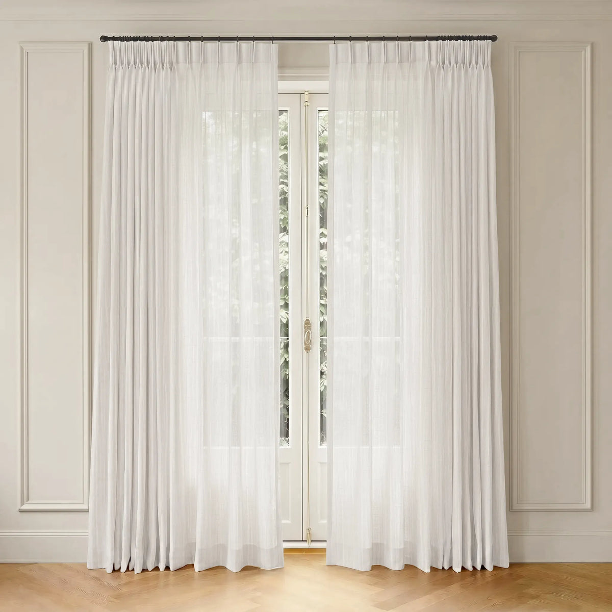 A pair of floor-length white sheer curtains hanging from a dark metal rod in a room with cream paneled walls and light wood flooring.