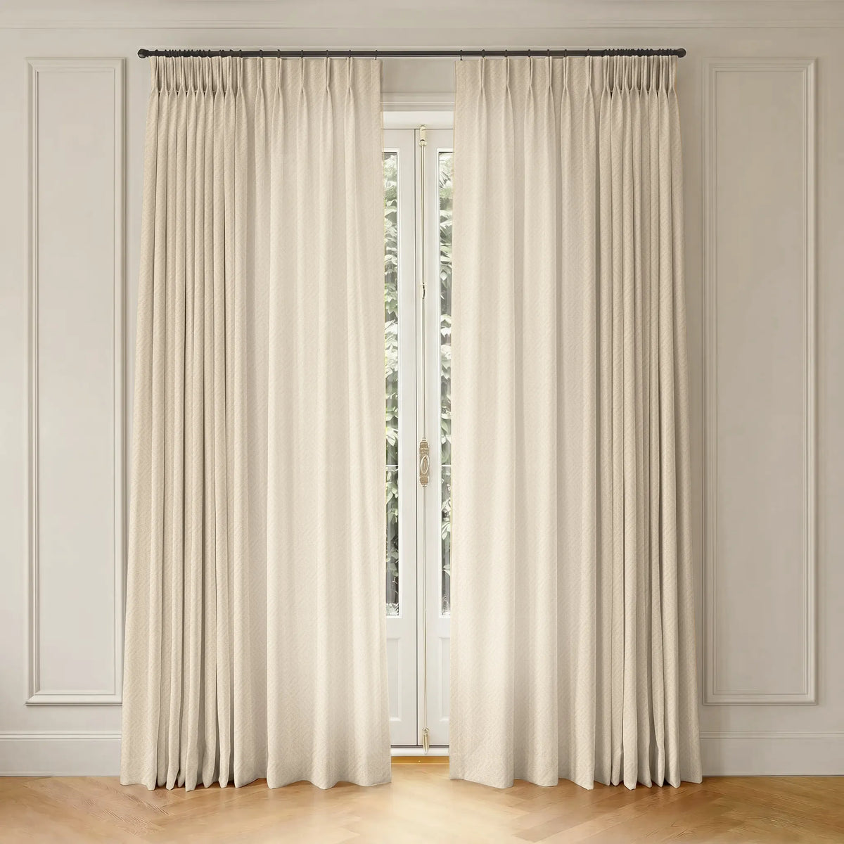 A pair of long cream-colored pleated window curtains hanging from a dark metal rod in a bright room with white paneled walls and light wood flooring.