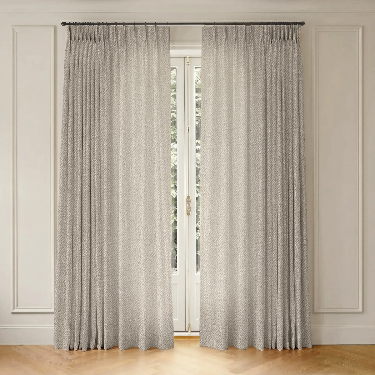 A pair of light gray pleated curtains hanging from a dark metal rod over a tall window, set against a cream paneled wall and light wood floor.