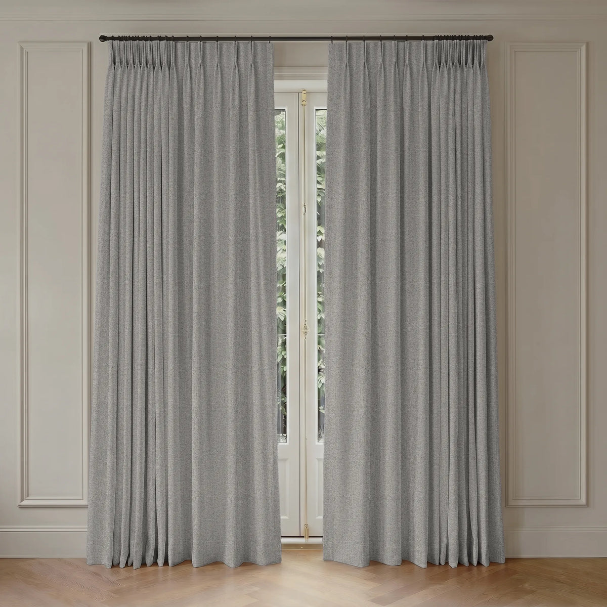 A pair of light gray pleated window curtains hanging from a dark metal rod in a room with cream paneled walls and a light wood floor.