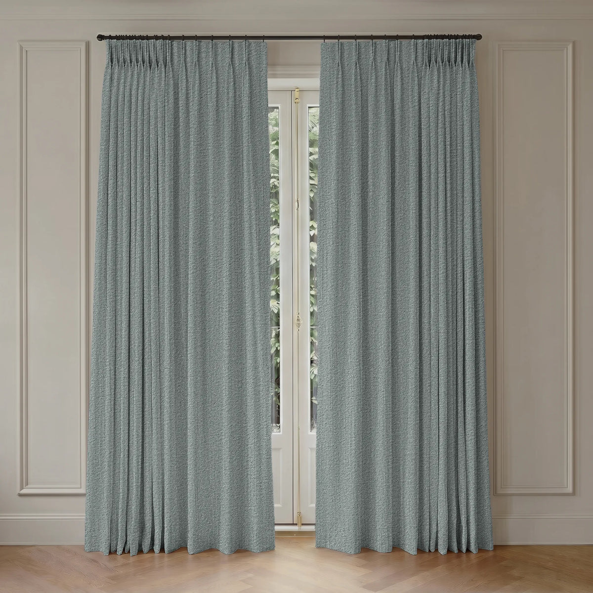 A pair of long pleated curtains in a dusty sage green color hanging from a black rod over a tall window in a room with beige paneled walls and light wood flooring.