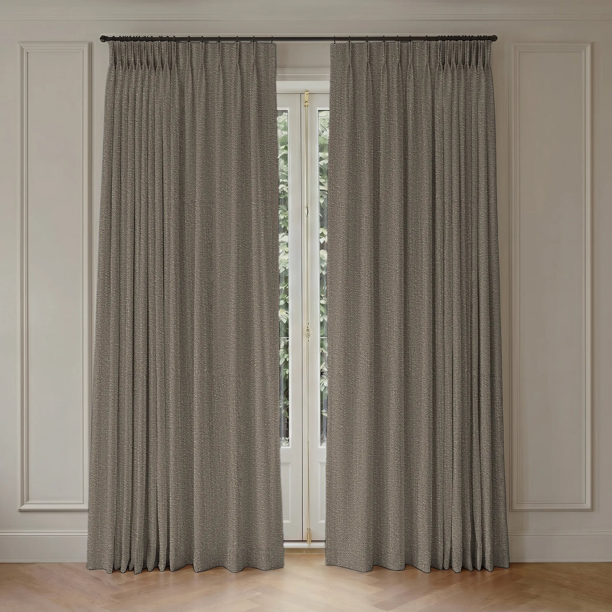 A pair of long pleated curtains in a textured taupe-gray fabric, hanging from a black rod over a tall window in a room with white paneled walls and light wood flooring.