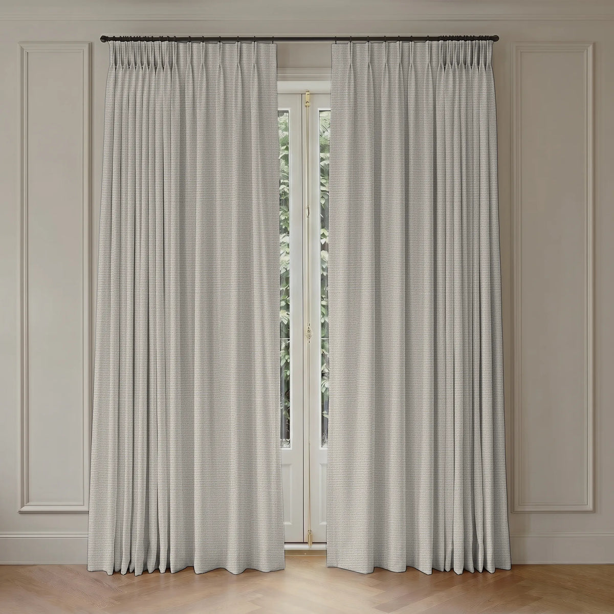 A pair of light gray pleated window curtains hanging on a dark metal rod in a room with cream paneled walls and a light wood floor.