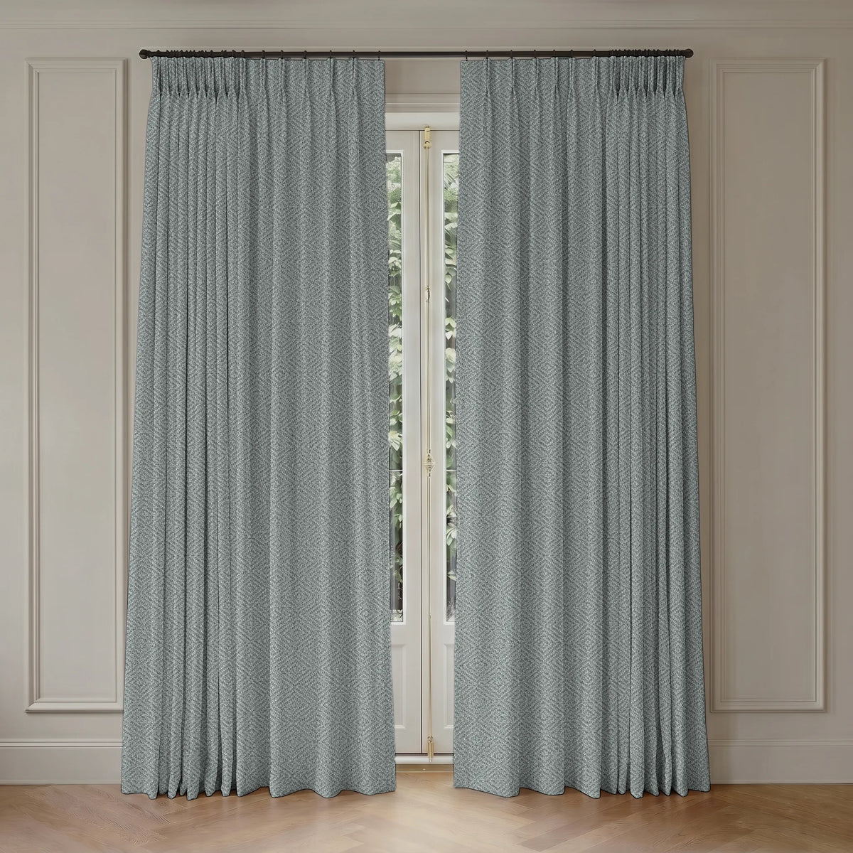 A pair of floor-length pleated curtains in a muted blue-gray color with a subtle repeating geometric pattern, hanging from a dark metal rod over a tall window in a room with cream paneled walls and light wood flooring.