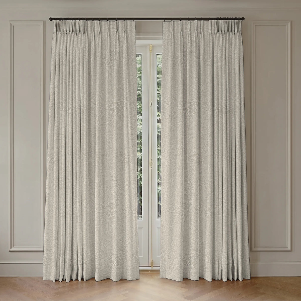 A pair of floor-length pleated curtains in a light beige textured fabric hanging on a dark metal rod over a tall window in a paneled cream room.
