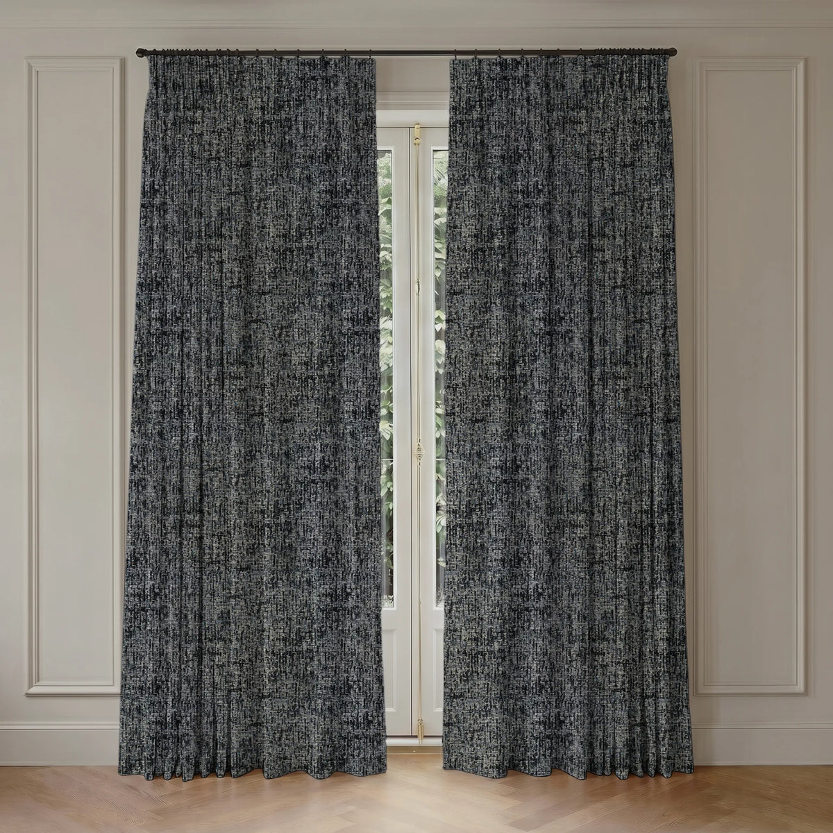 A pair of long window curtains with a textured, abstract black and gray pattern, hanging from a black rod in a room with cream paneled walls and a light wood floor.