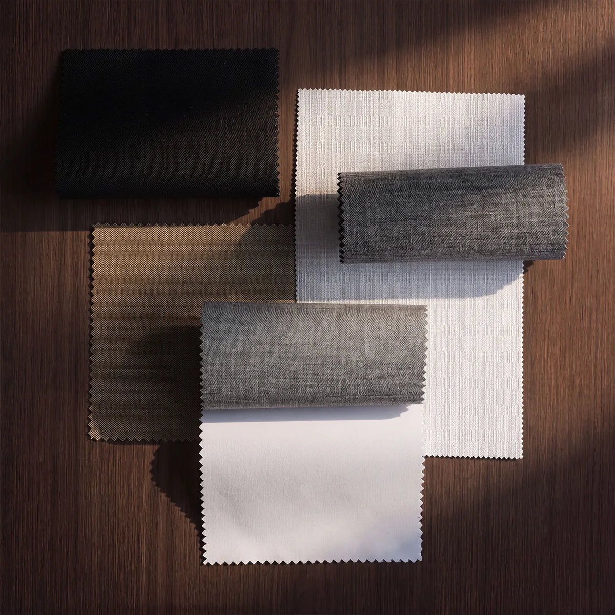 Four fabric swatch samples arranged on a dark wood surface, including black, taupe, and gray textured materials with some unfolded and some rolled sections.