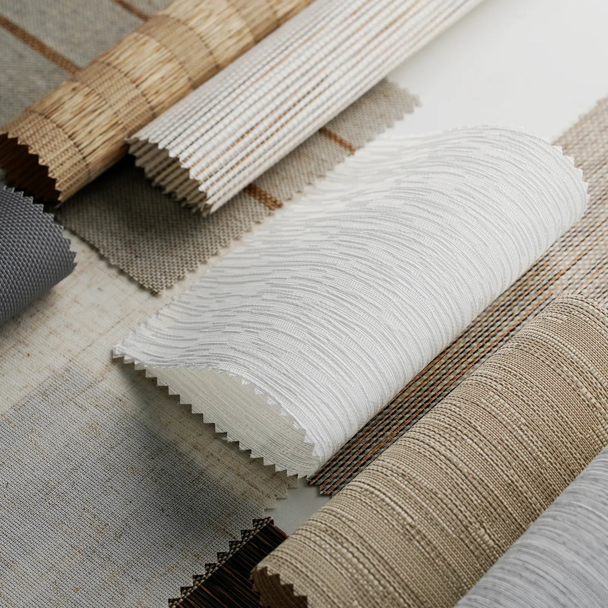 Close-up of rolled fabric or wallpaper samples in neutral tones, including white, beige, tan, and gray, arranged on a light surface.