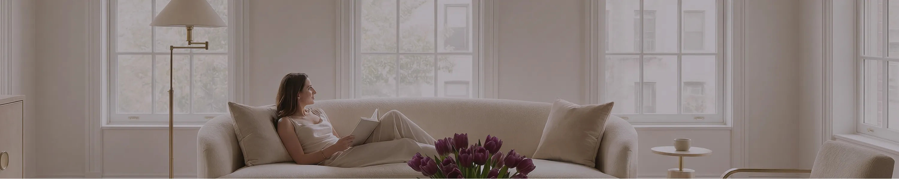 A woman relaxing on a beige sofa in a bright living room with large windows and soft natural light.