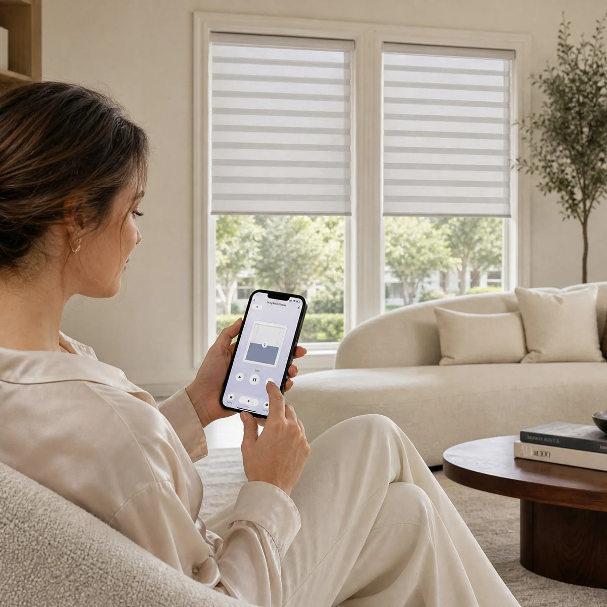 Woman using a smartphone app to control motorized light-filtering zebra blinds in a bright modern living room.