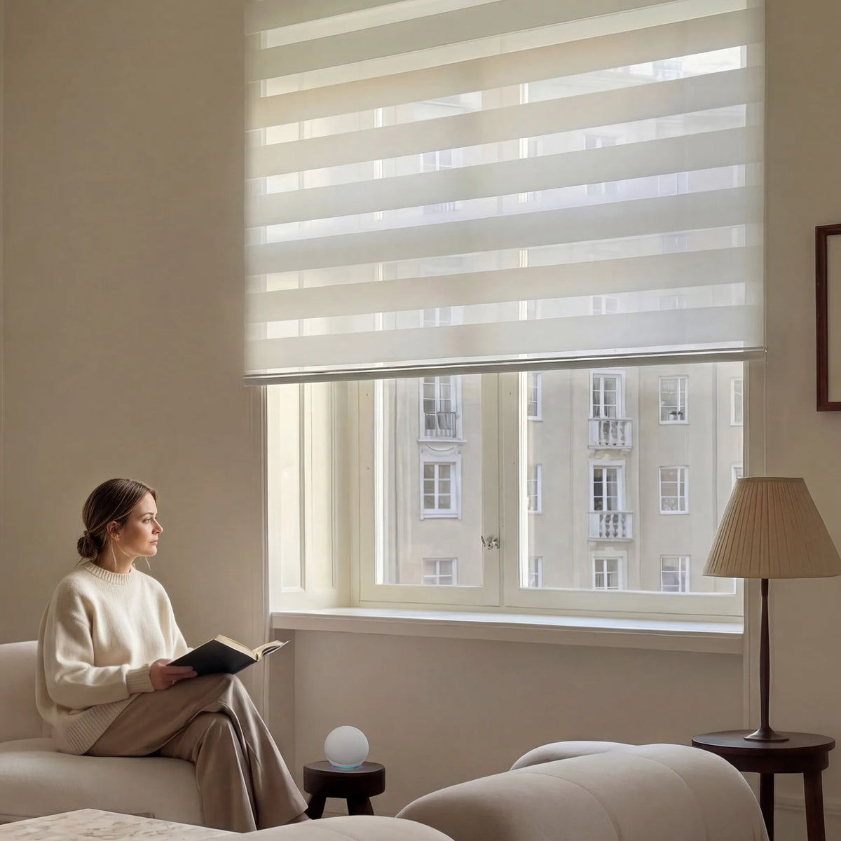 Woman reading on a beige sofa beside an off-white light-filtering zebra blind with a city view.