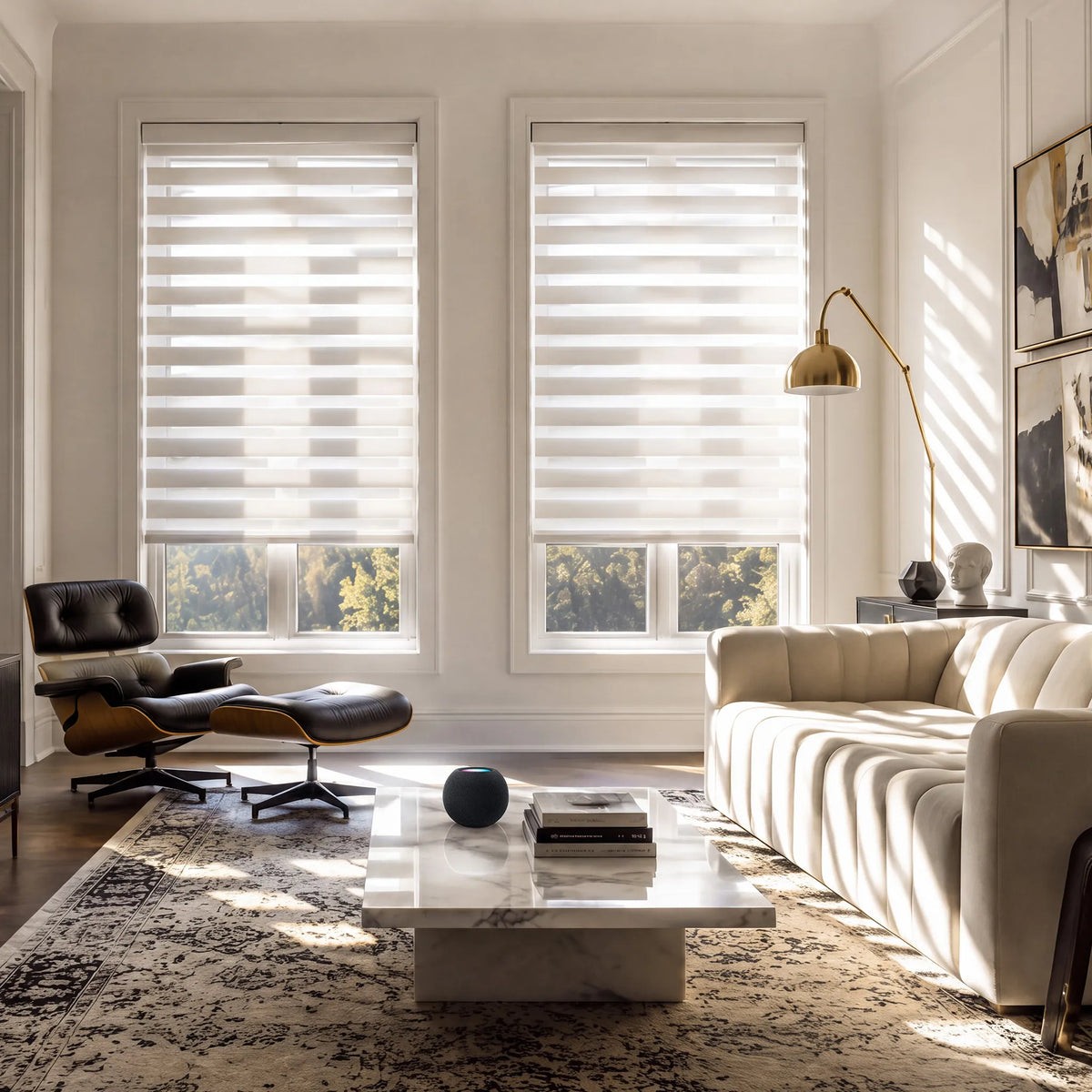Two white motorized light-filtering zebra blinds in a sunlit modern living room with a cream sofa and marble table.