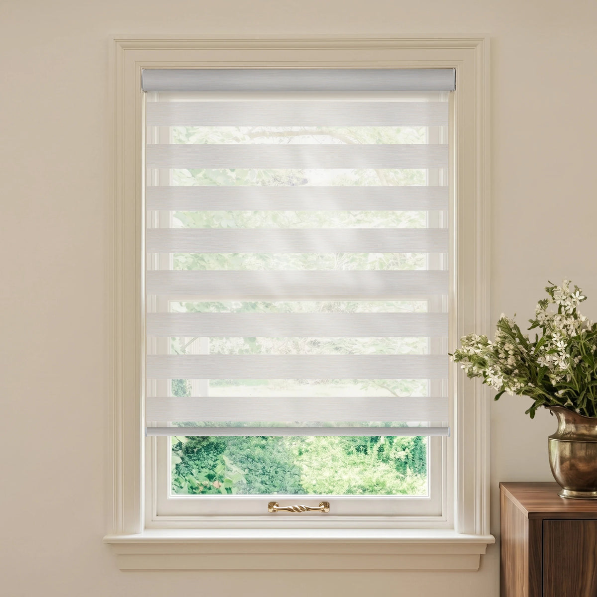 Linen Vanilla motorized light-filtering zebra blind with green trees outside and white flowers on a wooden table.