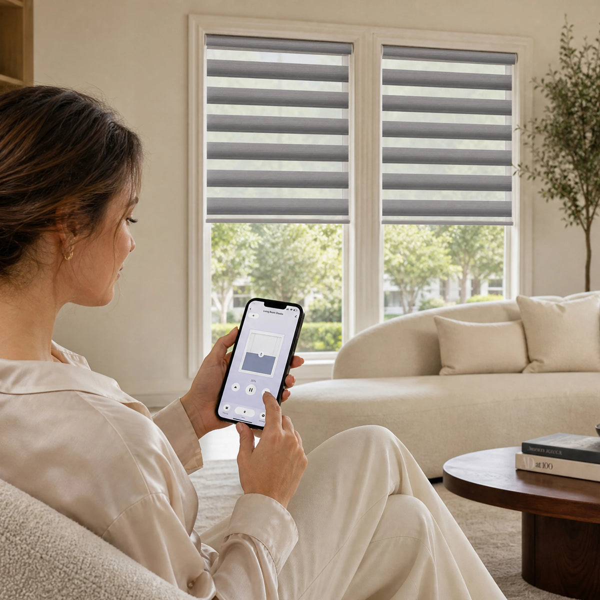 Woman using a smartphone app to control gray motorized room-darkening zebra blinds in a living room.