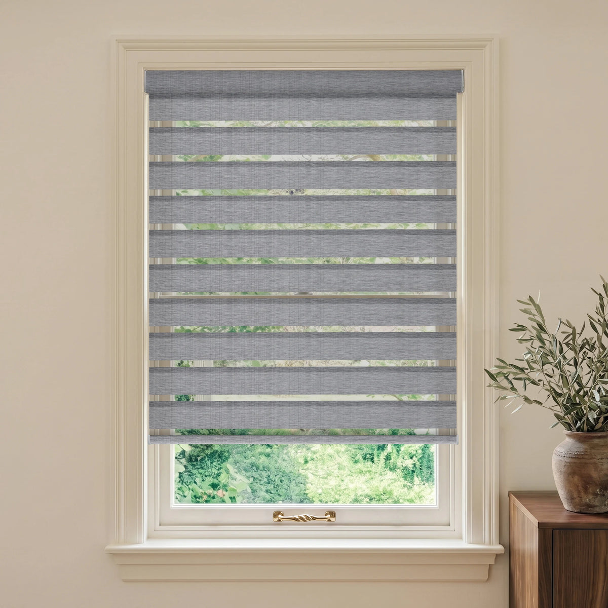 Grey motorized room-darkening zebra blind in a beige room beside a wooden table and olive branches.