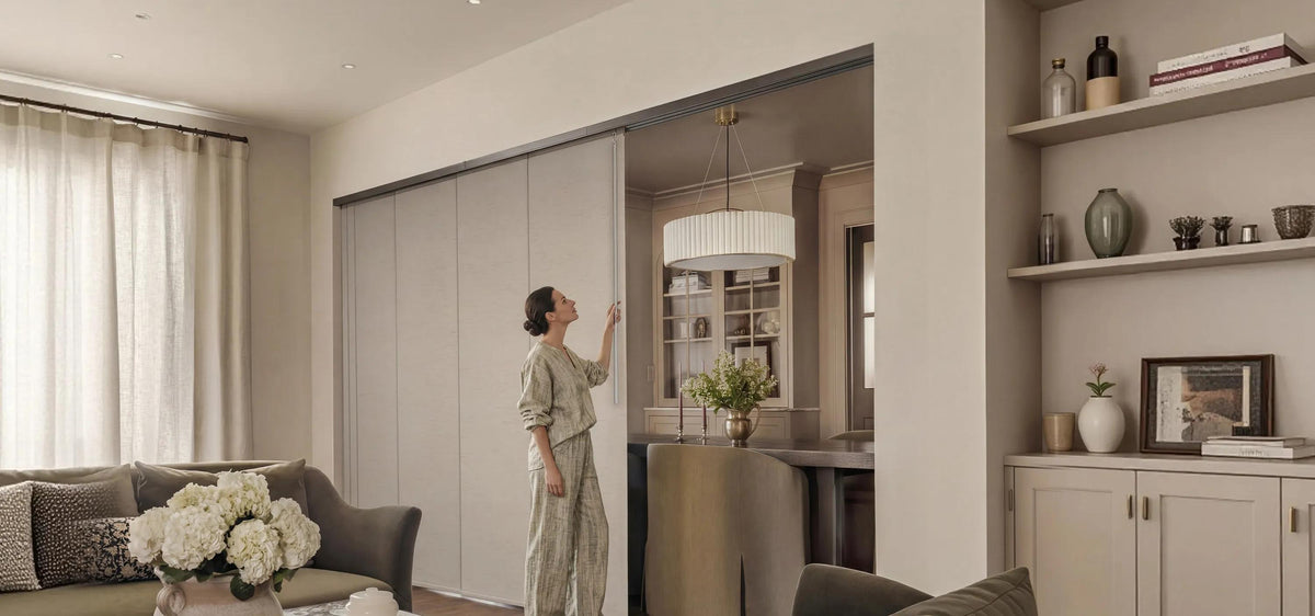 A woman adjusting sliding panel track blinds used as a chic room divider between a living area and dining space