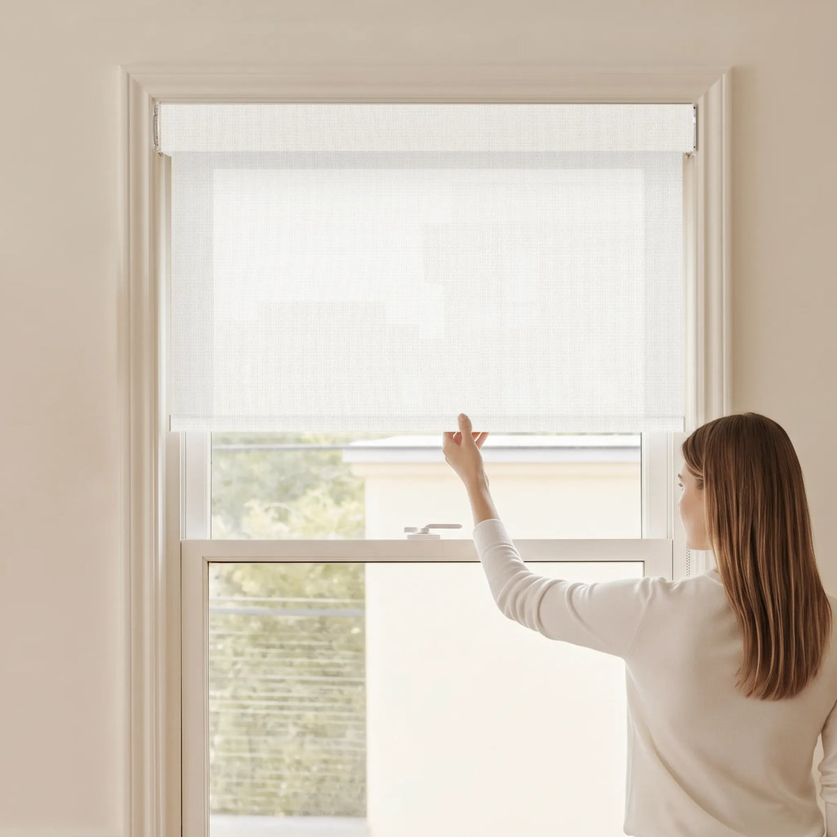 Woman adjusting a Graywind light filtering manual-motorized roller shade in Mist (White) fabric.