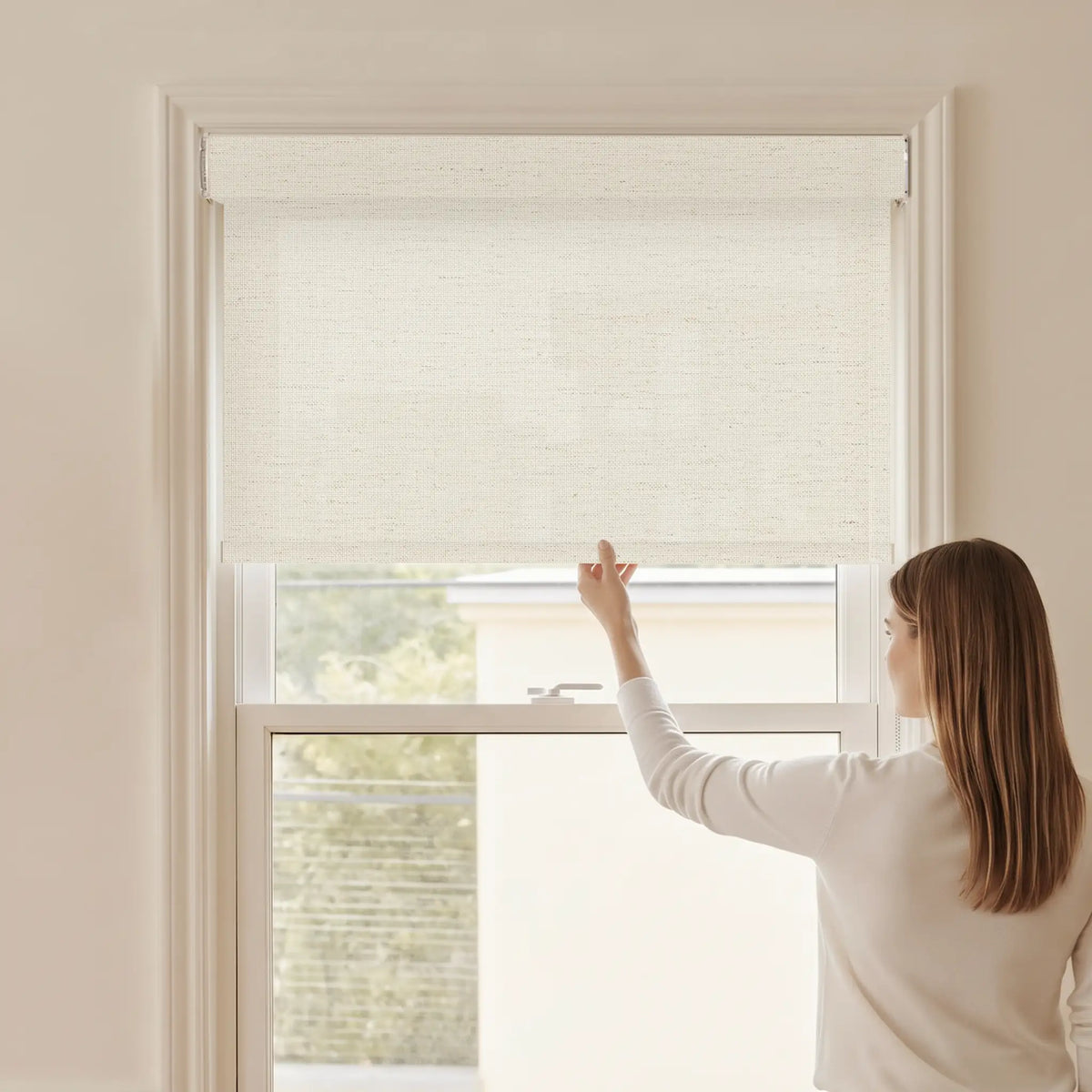 Woman adjusting a Graywind light filtering manual-motorized roller shade in Eden (Beige) fabric.