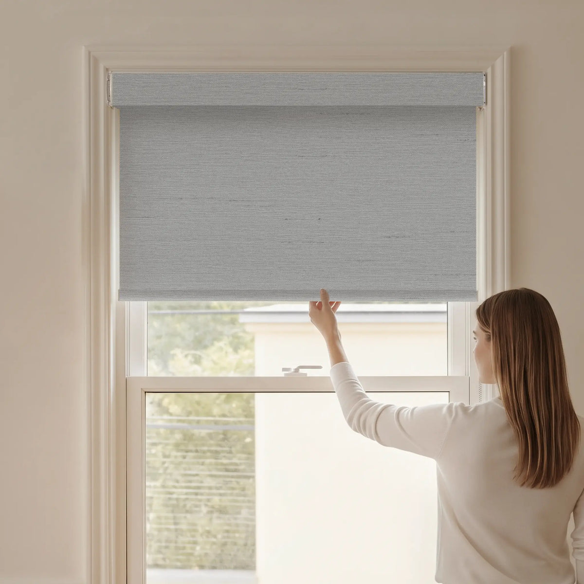 Woman adjusting a Graywind blackout manual-motorized roller shade in Horizon (Light Grey) fabric.