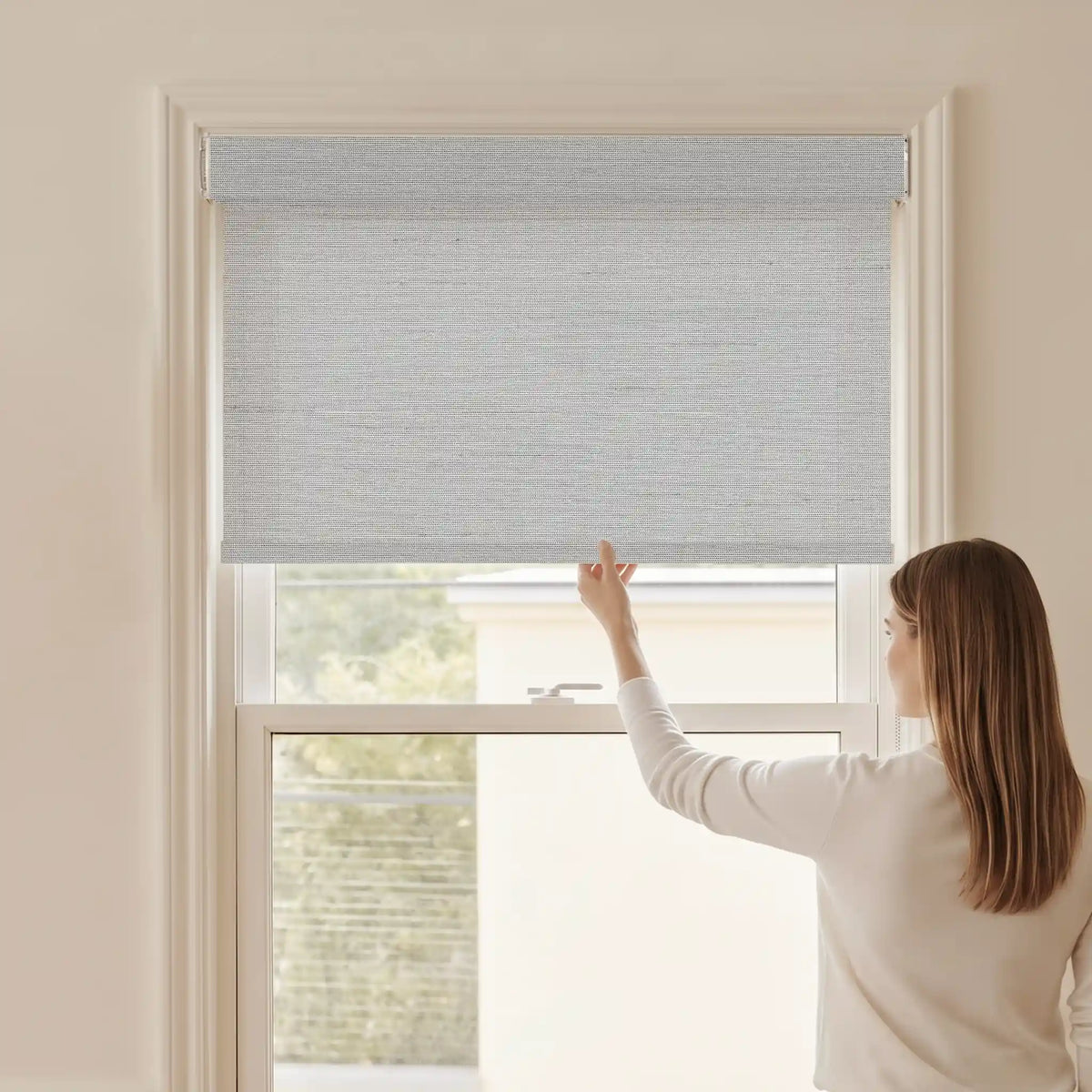 Woman adjusting a Graywind light filtering manual-motorized roller shade in Horizon (Light Grey) fabric.