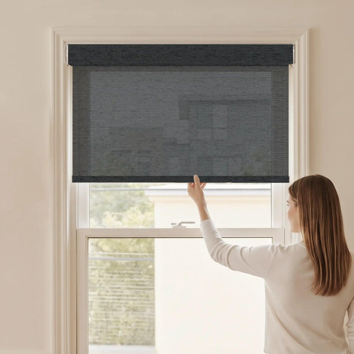 Woman adjusting a Graywind light filtering manual-motorized roller shade in Obsidian (Black) fabric.