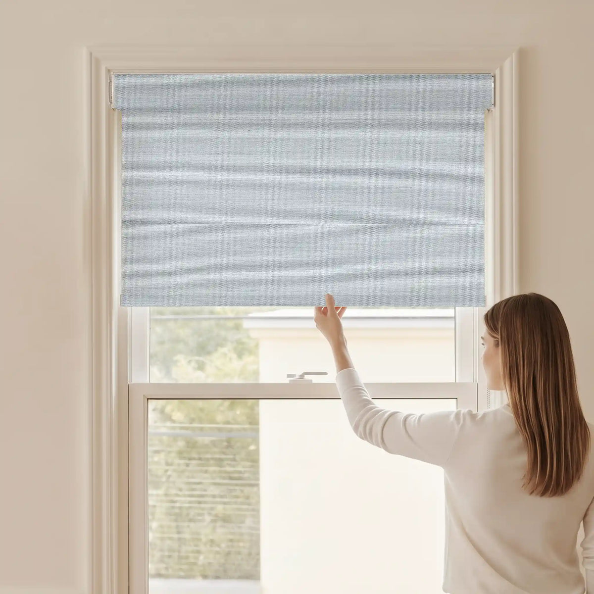 Woman adjusting a Graywind light filtering manual-motorized roller shade in Horizon (Sky Blue) fabric.
