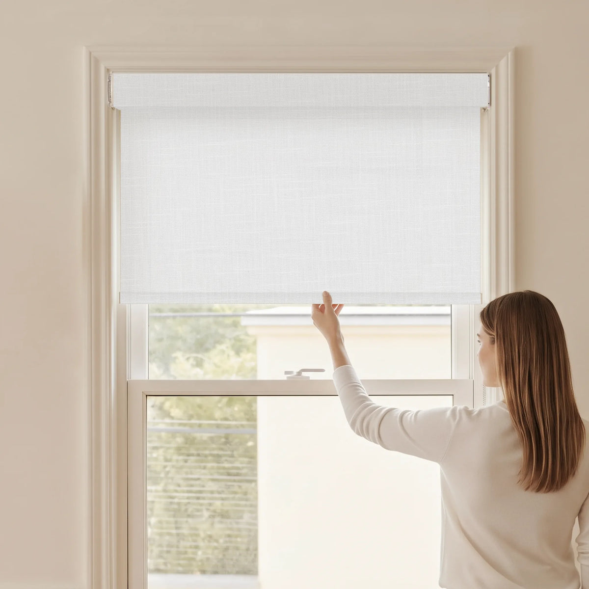 Woman adjusting a Graywind light filtering manual-motorized roller shade in Raw Rhythm (White) fabric.