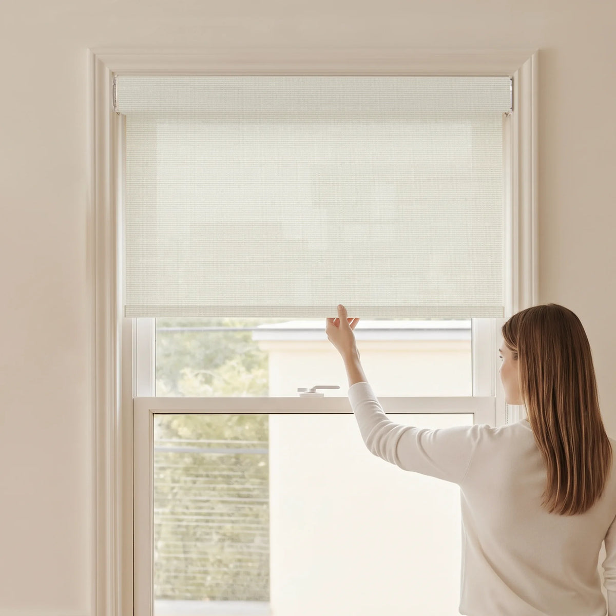 Woman adjusting a Graywind light filtering manual-motorized roller shade in Dune (Beige) fabric.