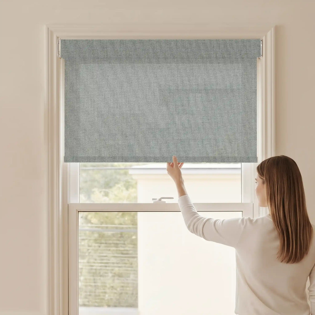Woman adjusting a Graywind light filtering manual-motorized roller shade in Dune (Smoke) fabric.