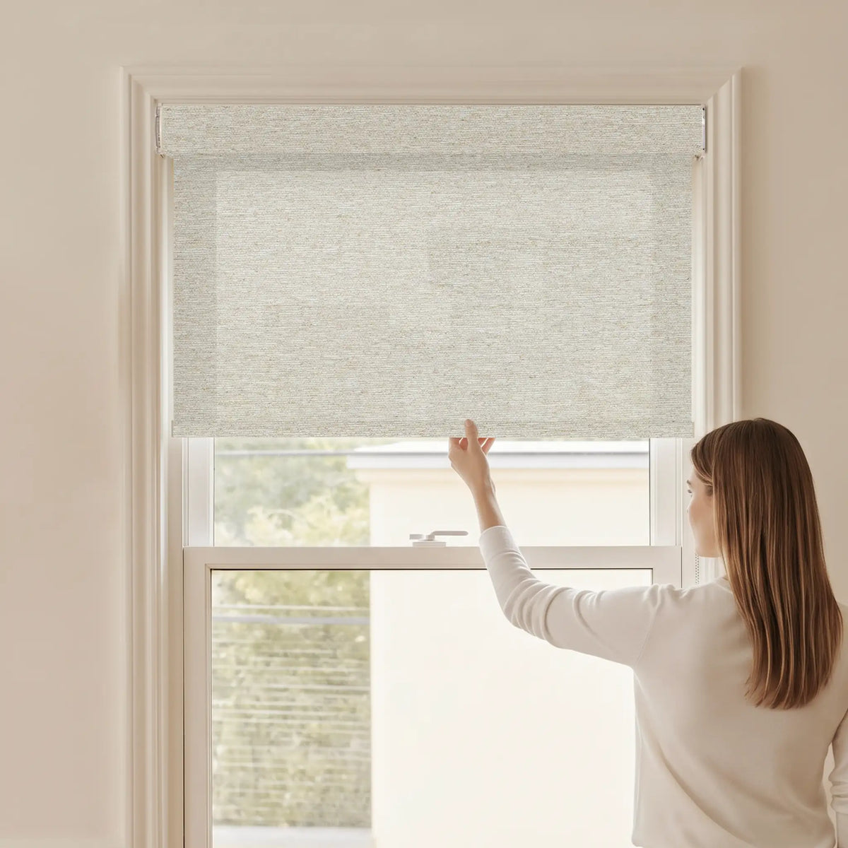 Woman adjusting a Graywind light filtering manual-motorized roller shade in Harvest (Bone) fabric.