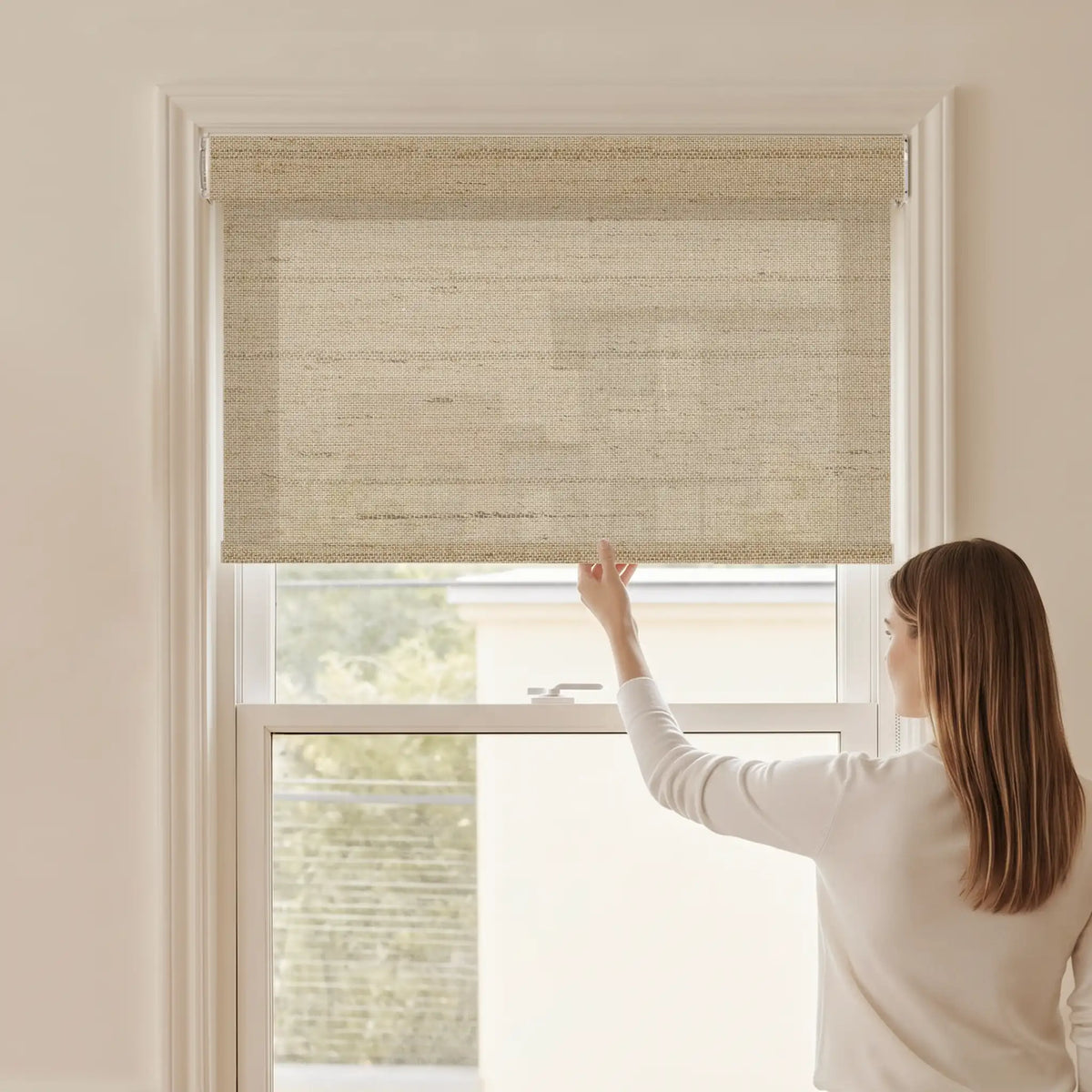 Woman adjusting a Graywind light filtering manual-motorized roller shade in Field (Camel) fabric.