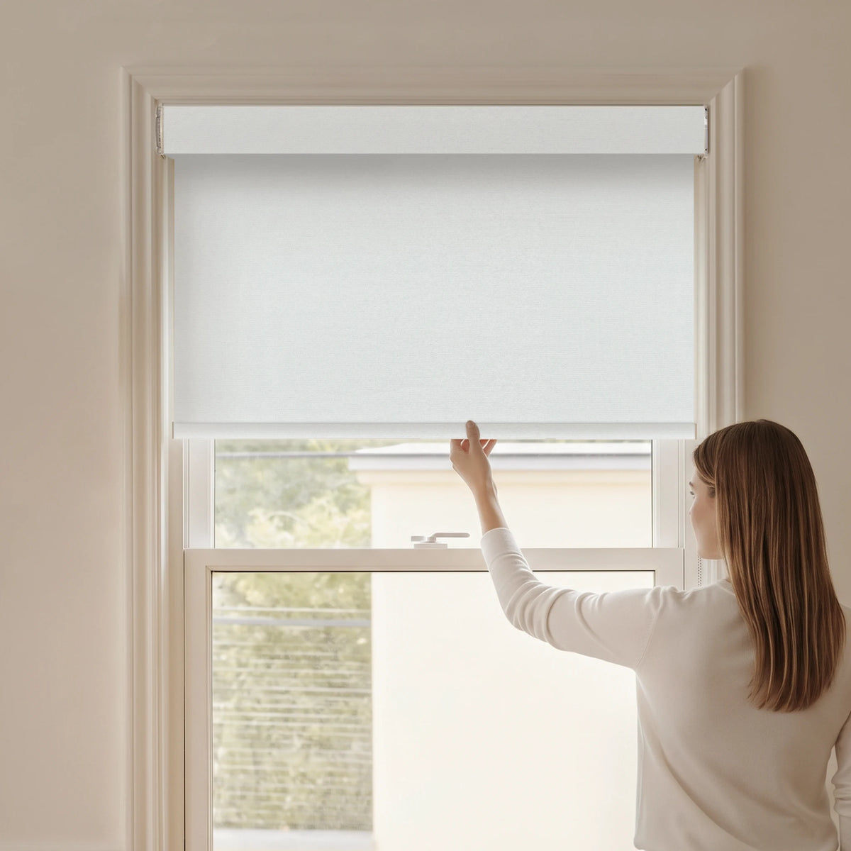 Woman adjusting a Graywind blackout manual-motorized roller shade in Horizon (White) fabric.