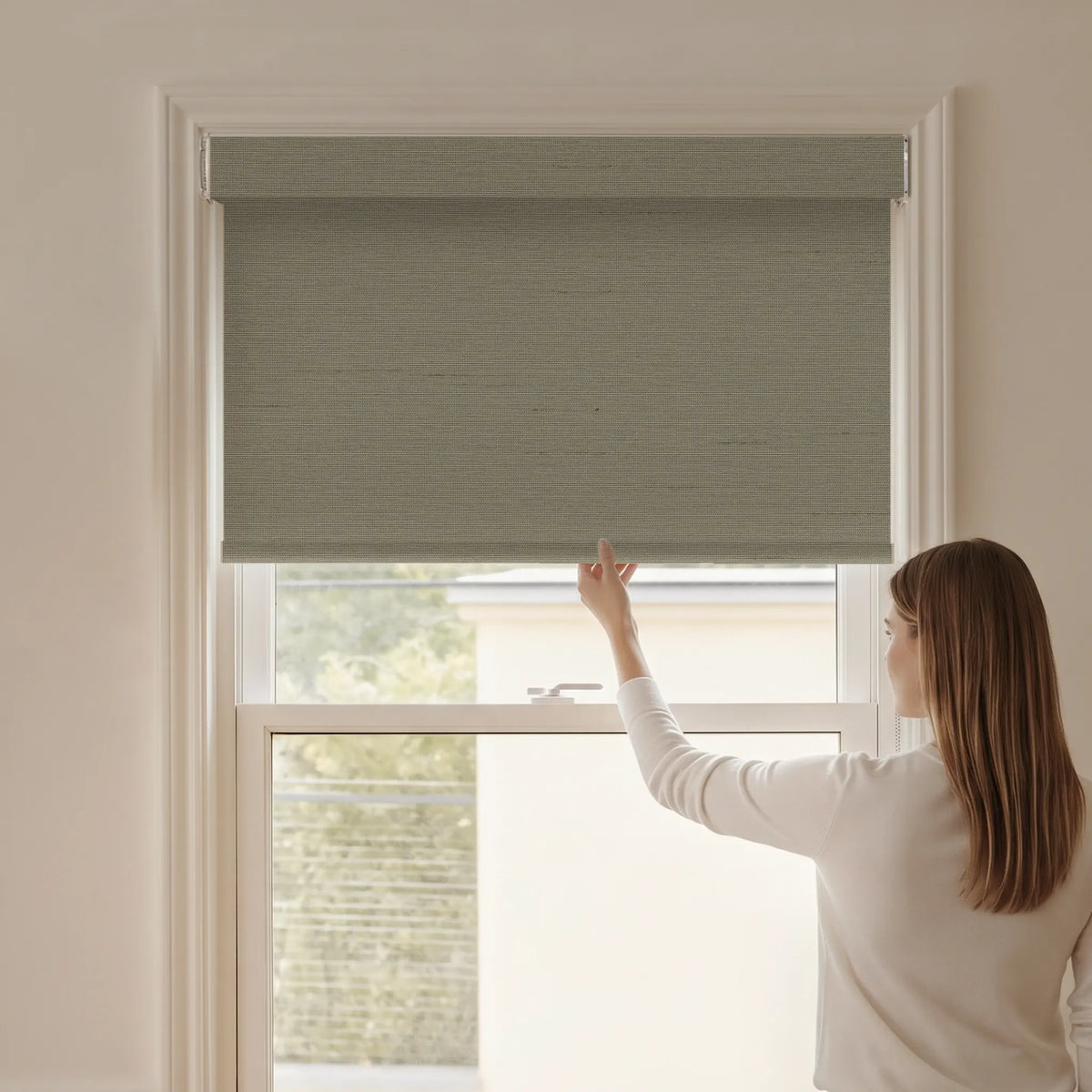 Woman adjusting a Graywind blackout manual-motorized roller shade in Horizon (Sand) fabric.
