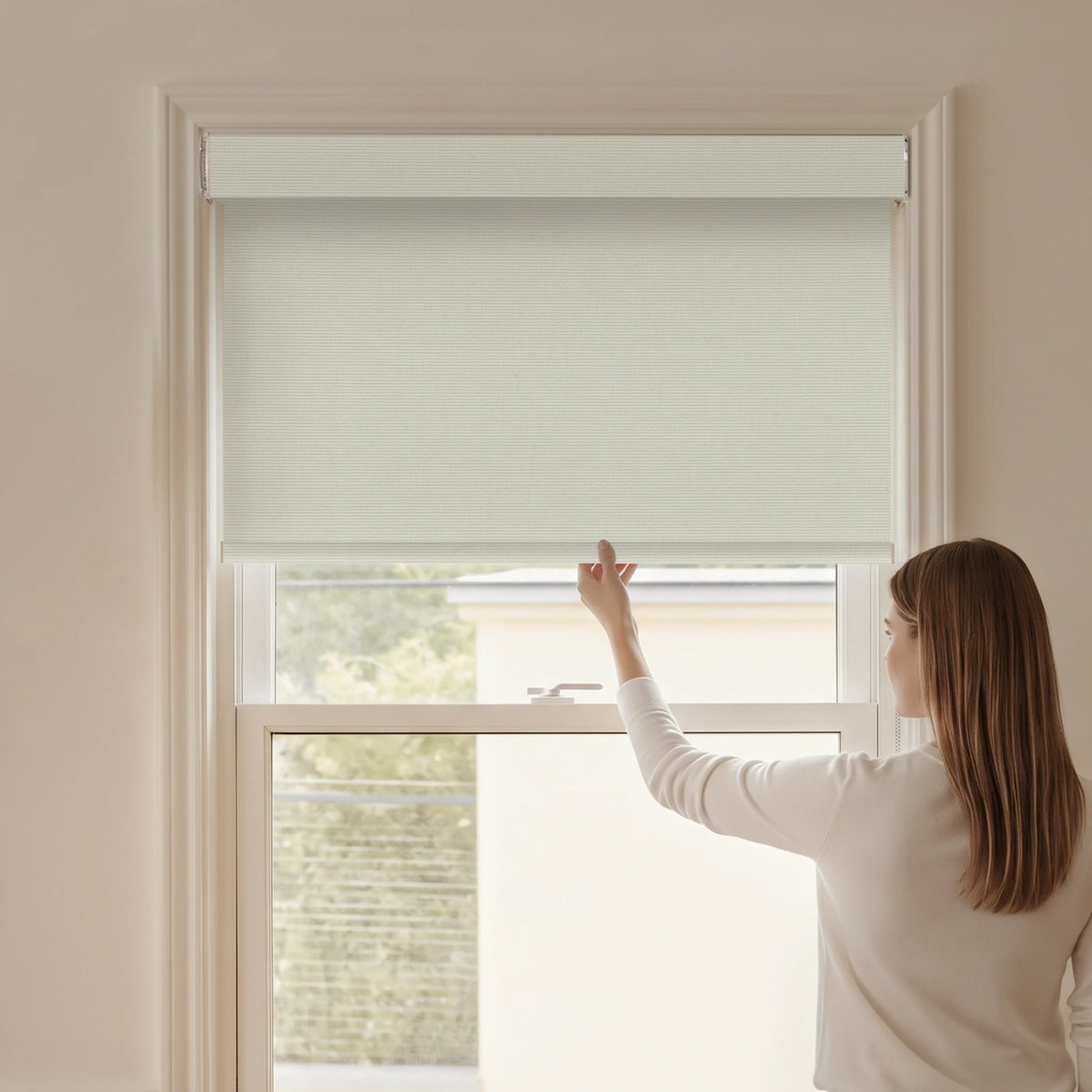 Woman adjusting a Graywind blackout manual-motorized roller shade in Dune (Beige) fabric.