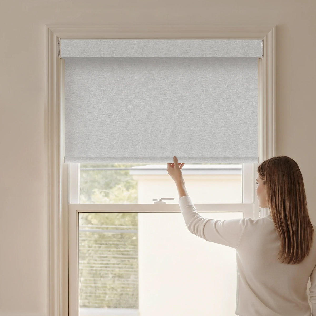 Woman adjusting a Graywind blackout manual-motorized roller shade in Plush Matte (White) fabric.