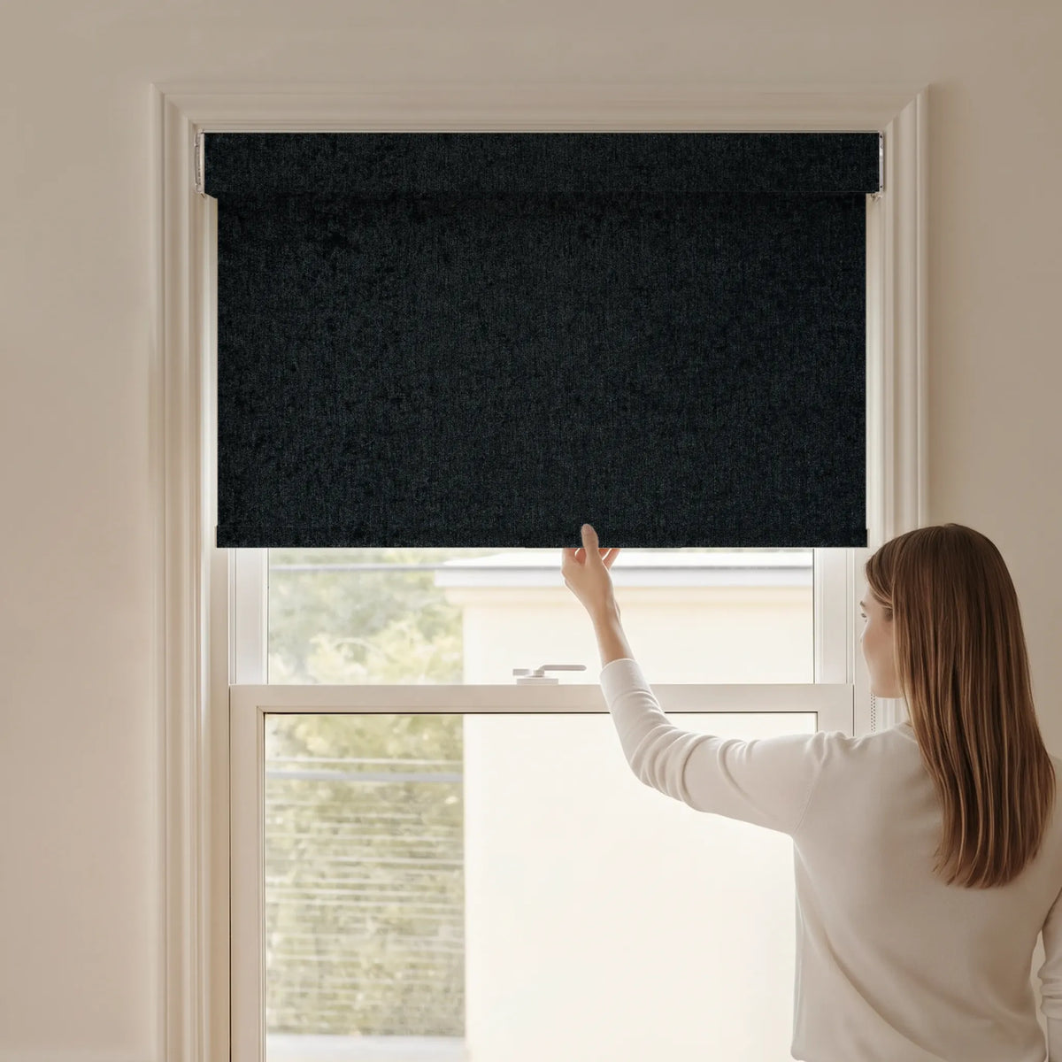 Woman adjusting a Graywind blackout manual-motorized roller shade in Plush Matte (Black) fabric.
