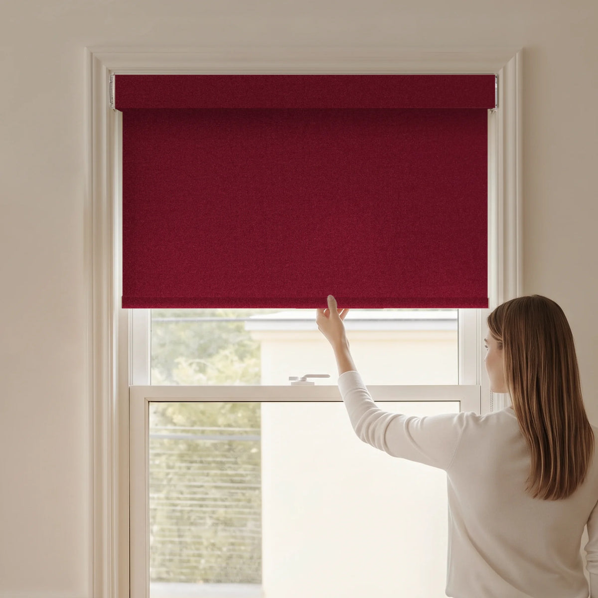Woman adjusting a Graywind blackout manual-motorized roller shade in Plush Matte (Wine) fabric.